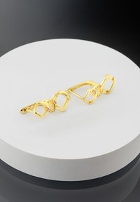 Armbanduhr in Goldoptik mit ineinander verwobenen quadratischen Formen, einem glänzenden Finish und dezenten Edelsteinakzenten, präsentiert auf einem runden weißen Stand.