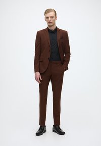 Ruskea puku, joka on valmistettu sileästä kankaasta ja yhdistetty mustaan paitaan. Slim fit -malli, yksimukinen takki ja mustat juhlakengät. Ei kuvioita tai koristeita.