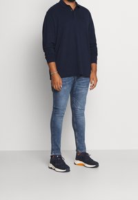 Polo à manches longues bleu marine, jeans ajustés bleu clair et baskets sombres avec des accents orange. Tissu lisse et design décontracté.