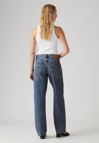 Modré džíny z denimu s volným střihem, viditelné švy na zadních kapsách a kožená nášivka na pasu. Stylizováno s bílým tričkem s úzkými ramínky.