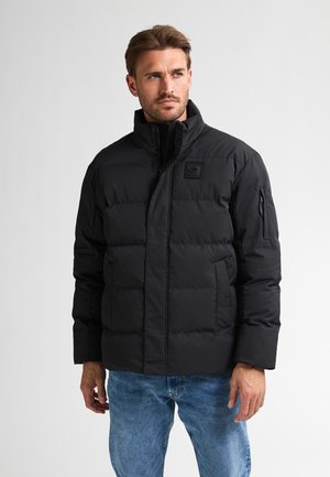 Man met kort bruin haar, gekleed in een zwarte pufferjack en een blauwe spijkerbroek, die iets naar rechts kijkt tegen een effen lichte achtergrond.
