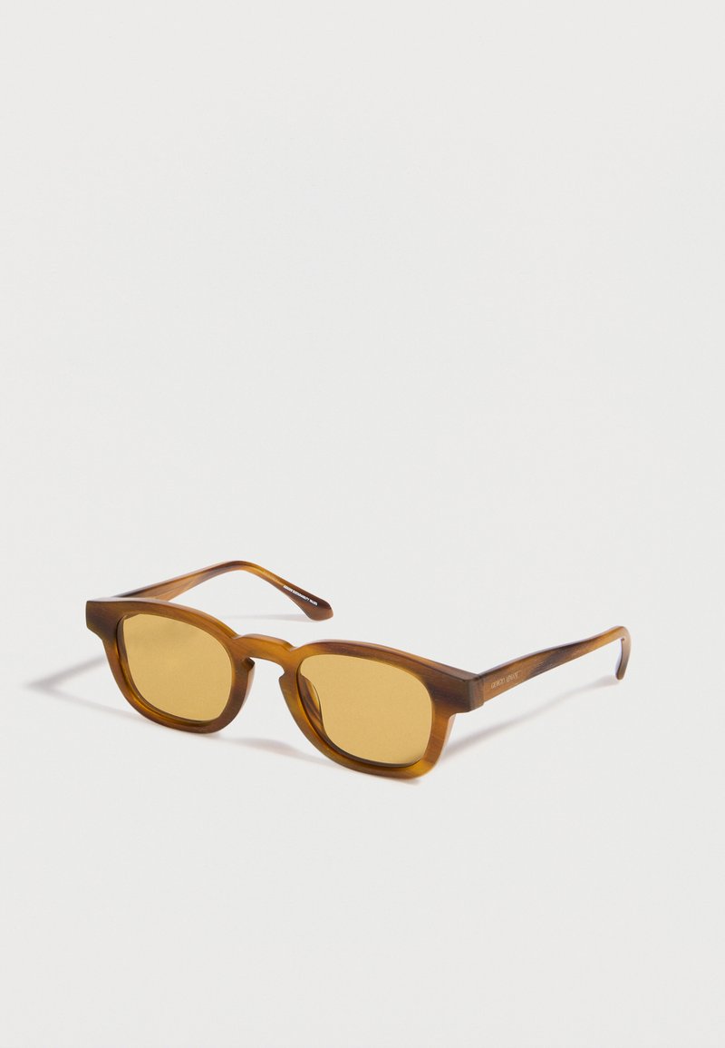 Lunettes de soleil rectangulaires translucides marron avec des verres ambrés posées sur une surface blanche, inclinées vers la droite.