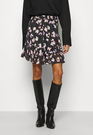 Woman wearing a black long-sleeve top, black pleated floral mini skirt with pink and white spots, and black knee-high heeled boots.