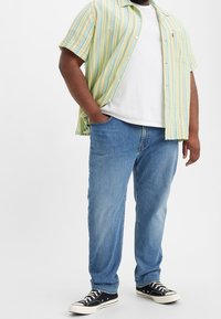 Man som bär blå jeans, svarta kanvas sneakers, vit t-shirt och en oformad kortärmad skjorta med gula ränder, med händerna delvis i fickorna.