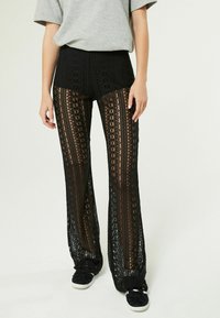 Pantaloni in pizzo nero con vita alta, motivi intricati, gambe svasate e abbinati a scarpe nere. Il tessuto è trasparente con dettagli di design visibili.