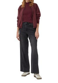 Vrouw in een bordeauxrode jack met ritssluiting, bijpassend bordeaux top, zwarte wijde jeans en beige sneakers op een witte achtergrond.