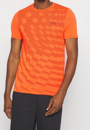 Man draagt een feloranje sportshirt met korte mouwen en een patroonstructuur, en zwarte shorts, staand tegen een effen achtergrond.