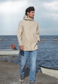 Beige vattentät jacka med huva, med en mjuk yta, dragkedjestängning och sidofickor, kombinerad med blå jeans och grå sneakers.