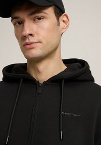 Czarna bluza rozpinana z kapturem wyłożonym materiałem, z praktycznymi sznurkami i małym logo na piersi. Gładka, miękka tekstura materiału.