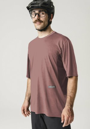 Mann trägt einen schwarzen Fahrradhelm, eine Brille, ein mauvefarbenes kurzärmeliges Hemd und schwarze Hosen, steht vor einem schlichten, hellen Hintergrund.