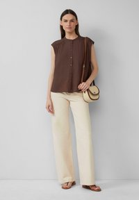 Bruine blouse met korte mouwen, knoopsluiting en geplooide schouderdetails, gedragen met lichtbeige wijde broek en bruine sandalen.