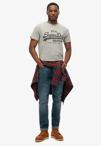 Camiseta gris con estampado negro, pantalones vaqueros azules, camisa de cuadros roja y verde atada a la cintura, botas de cuero marrón con suela blanca.
