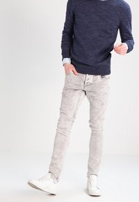 Pull en maille bleu marine, chemise à col bleu clair en dessous, associé à un jean skinny gris clair déchiré et des baskets blanches.