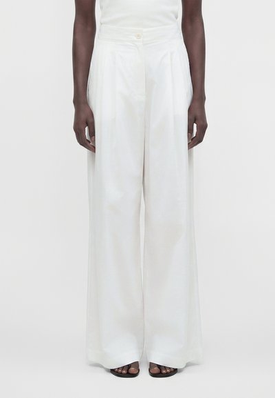 Modèle portant un pantalon blanc taille haute à jambes larges avec des plis, associé à des sandales noires à bouts ouverts sur un fond neutre.