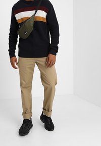 Marinblå stickad tröja med röd, vit och gul rand, khakifärgade upprullade byxor, svarta sneakers och en grön axelremsväska.