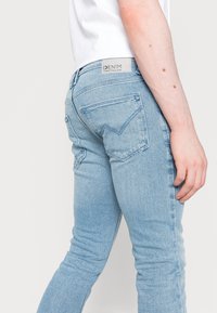 Osoba oblečená v světlých modrých džínách a bílém tričku, zobrazující zadní a boční pohled na džíny s viditelnými zadními kapsami.