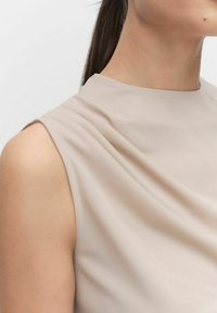 Beige ærmeløs top med draperet halsudskæring, blødt stof, glat tekstur og minimalistisk design. Beklædningsgenstanden har sarte folder ved skulderen.