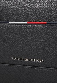 Sac en cuir noir texturé avec un logo subtil indiquant "TOMMY HILFIGER" en argent métallique. Doté d'accents rayés rouges, blancs et bleus.