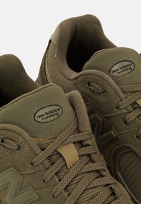 Para oliwkowo-zielonych butów do biegania marki New Balance z zamszową i siateczkową tkaniną, sznurowanych i prezentujących logo marki na języku i boku.