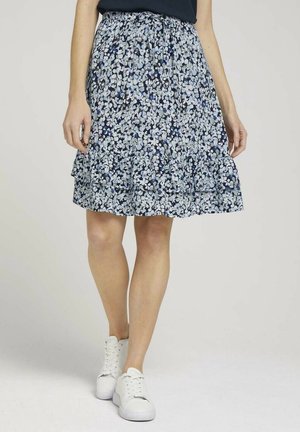 Woman wearing a blue floral knee-length skirt, black top, and white sneakers, standing with legs slightly crossed.