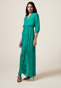 Rochie maxi în nuanță de teal, cu decolteu adânc în V, mâneci bufante, un detaliu plisat în față și material fluid. Pur ati cu sandale negre cu toc.