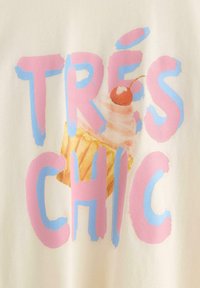 Krämfärgad skjorta med en cupcake-grafik med rosa glasyr och ett körsbär. Fet rosa och blå text säger "TRÈS CHIC."