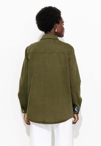 Une personne avec des cheveux courts et bouclés porte une chemise à manches longues olive et un pantalon blanc, de dos, devant un fond blanc uni.