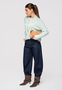 Femme avec queue de cheval portant un cardigan bleu clair, un jean large foncé, des bottes en suède marron, tenant une pochette en suède beige.