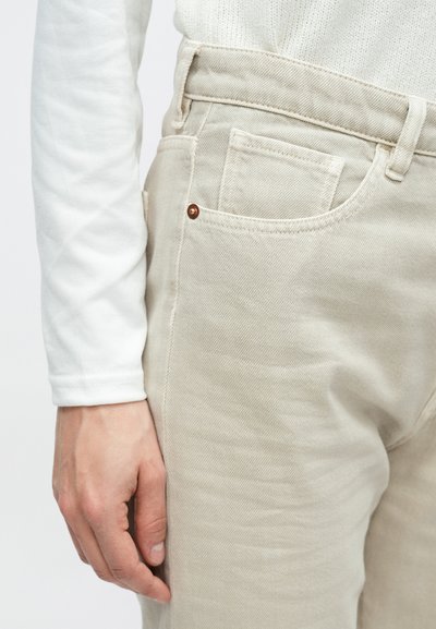 Μπεζ παντελόνι τζιν με υφή, μπροστινή τσέπη με ραφές, λεπτομέρεια από χαλκόβαφτα καρφιά και φορεμένο με λευκό πουκάμισο μακρυμάνικο.