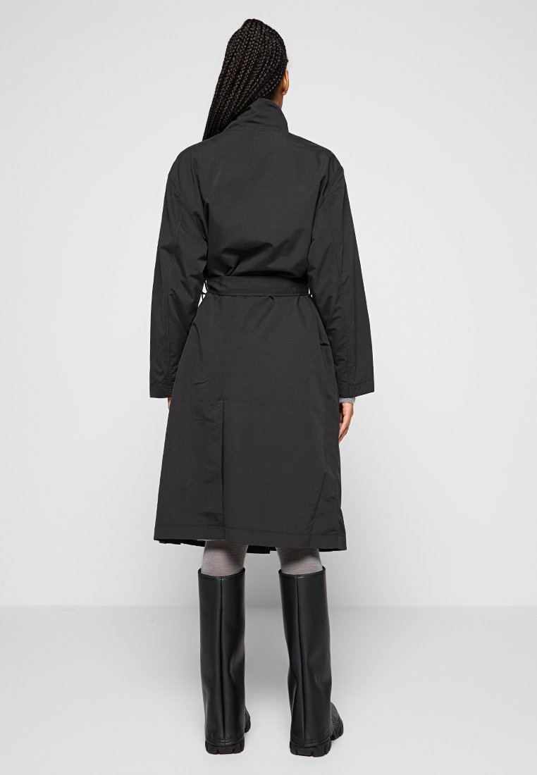 Schwarzer Langarmmantel mit tailliertem Gurt, knielangem Saum und hohem Kragen. Glatte Stoffstruktur, minimalistisches Design, keine sichtbaren Muster.