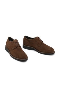 Chaussures en suede marron pour hommes, à lacets, avec des semelles en caoutchouc noires, présentées côte à côte sur un fond blanc.
