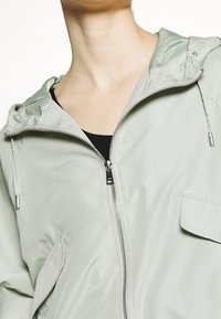 Jasnozielona kurtka z zamkiem i kapturem, wykonana z gładkiego materiału, z kieszenią z przodu i srebrnymi detalami zamka.