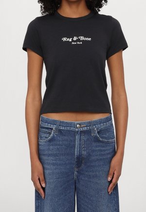 Femme portant un t-shirt court noir Rag & Bone et un jean en denim bleu taille haute, debout les bras relâchés le long du corps.