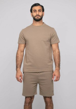 Homme debout face à l'avant, portant une chemise et un short beige texturés assortis à manches courtes, sur un fond clair uni.