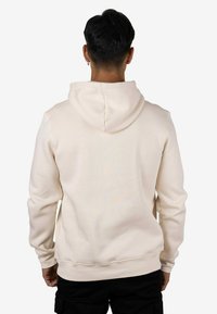 Sudadera con capucha de color crema y textura suave, con capucha ajustable mediante cordón y puños acanalados, vista desde atrás, usada sobre pantalones oscuros.