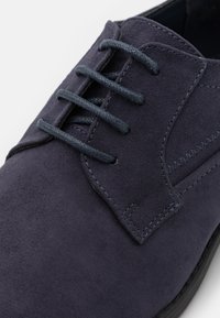 Chaussure en daim bleu marine avec une texture lisse, dotée de lacets ton sur ton, d'une couture décorative près de l'ouverture et d'un design à bout rond.