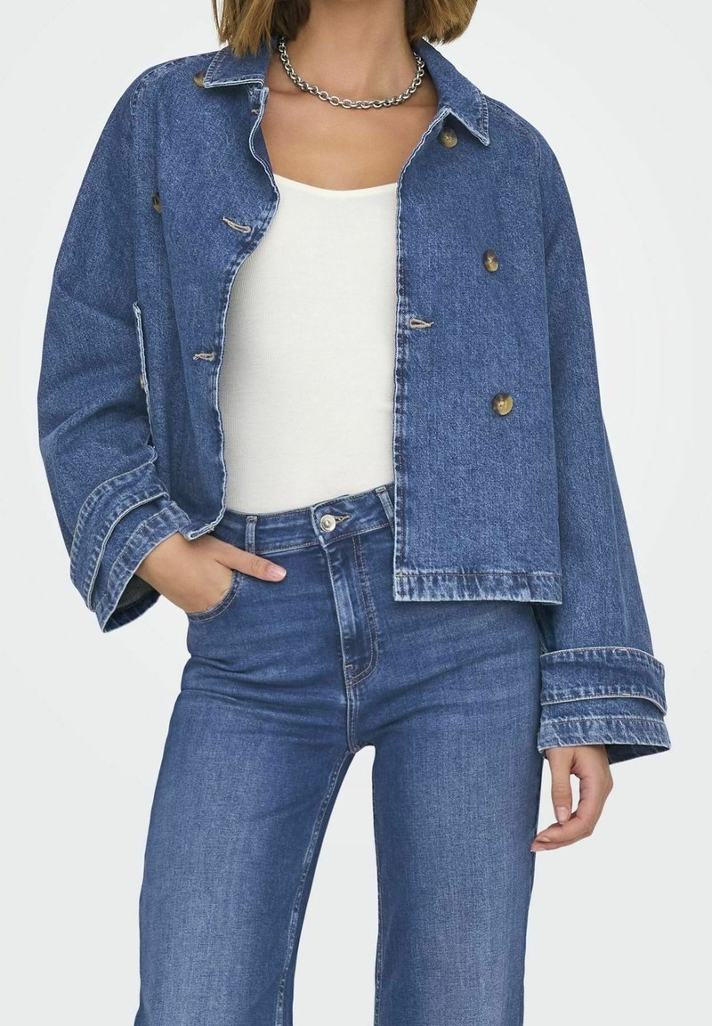 Modrá džínová bunda s krátkým střihem, velkými knoflíky a širokými manžetami, nošená přes bílé tričko a doplněná modrými džínami.