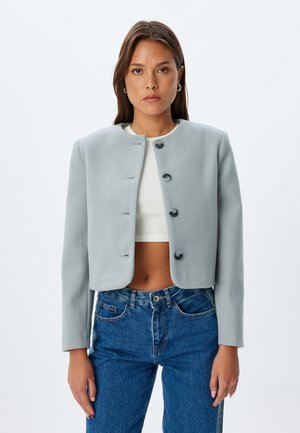 Chaqueta gris corta con cuello redondo, cinco cierres de botón negro, textura suave y mangas largas, combinada con una blusa blanca y jeans azules.