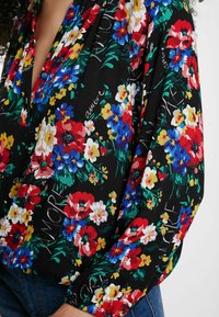 Schwarze Bluse mit lebhaftem roten, blauen, gelben und weißen Blumenmuster sowie subtiler Schrift, getragen mit blauen Jeans.