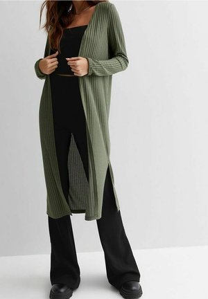 Kobieta w otwartym, prążkowanym, oliwkowozielonym kardiganie nałożonym na czarny top bandeau i szerokie czarne spodnie, stojąca na tle gładkiej ściany.