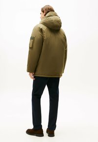 Doudoune verte olive avec capuche, poignets élastiques et une poche zippée sur la manche gauche, portée avec un pantalon bleu marine et des chaussures marron.