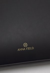 Surface en cuir noir avec logo et emblème ANNA FIELD dorés embossés près du bas au centre.