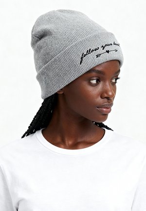 Gray knit beanie with a ribbed texture features black embroidered text "follow your heart" and a heart icon. White shirt worn underneath.