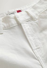 Valkoiset denimmekot, joissa on sileä pinta, vyölenkit ja vyötärönauhan sisäpuolella oleva punaisin yksityiskohdin varustettu etiketti. Lähikuva kankaan yksityiskohdasta.