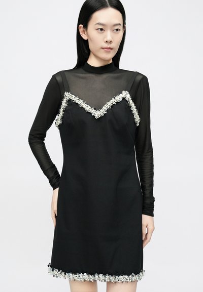 Melna kleita ar caurspīdīgu garām piedurknēm un V veida izgriezumu, malas un apakša izrotāta ar ziedu aplikācijām.