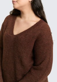 Brązowy sweter dzianinowy z wycięciem w serek, o luźnym kroju i prążkowanym kołnierzu. Tekstura wygląda na miękką i lekko puszystą.