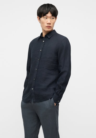 Νεαρός με κοντά μαύρα μαλλιά, που φοράει ένα σκούρο πουκάμισο με μακριά μανίκια και γκρι παντελόνι, στέκεται με τα χέρια στις τσέπες.