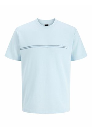 Niño de pie de frente con camiseta azul clara, vaqueros anchos grises y zapatillas blancas sobre un fondo claro y liso.