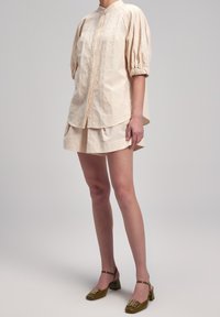 Femme portant une tenue beige texturée composée de deux pièces avec des manches bouffantes et un short assorti, associée à des chaussures à talons bas vert olive.