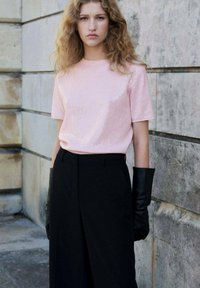 Joven mujer con cabello rizado, vestida con una blusa de manga corta color rosa claro, pantalones negros de talle alto y guantes negros largos, de pie junto a una pared de piedra.